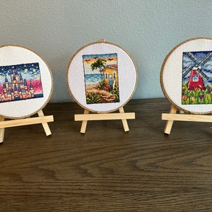 May include: Three framed cross-stitch embroidery pieces on wooden easels. The first piece depicts a colorful castle with stars in the sky. The second piece shows a yellow beach house with a palm tree and ocean waves. The third piece features a red windmill with flowers in a field.
