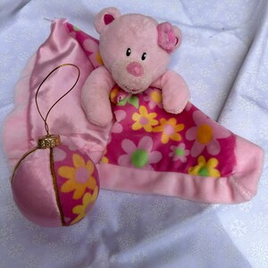 Op de afbeelding: Roze pluche teddybeer met een roze bloem op zijn oor, genesteld in een roze deken met bloemenpatroon. Een roze en gele ornament met een gouden rand is ook inbegrepen.