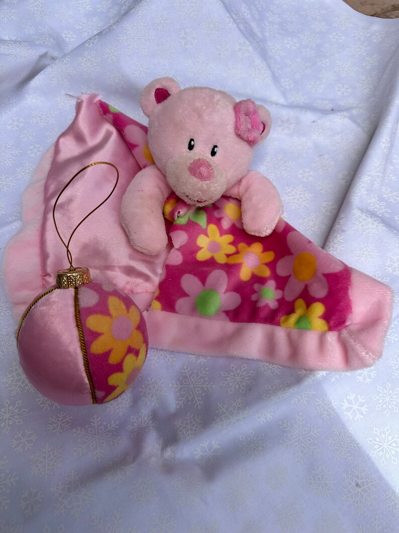 Op de afbeelding: Roze pluche teddybeer met een roze bloem op zijn oor, genesteld in een roze deken met bloemenpatroon. Een roze en gele ornament met een gouden rand is ook inbegrepen.