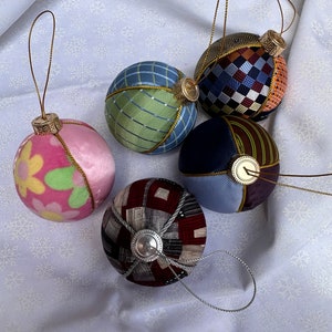 Op de afbeelding: Vijf handgemaakte kerstballen met verschillende patronen en kleuren. De ballen zijn gemaakt van stof en hebben een gouden rand. Ze hangen aan een witte achtergrond.