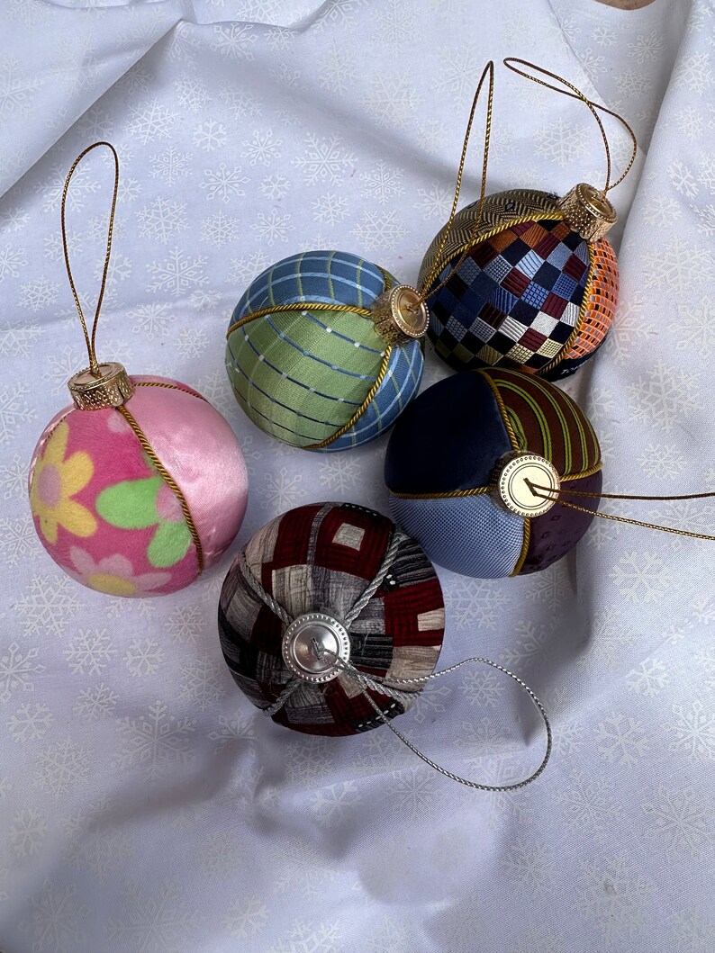 Op de afbeelding: Vijf handgemaakte kerstballen met verschillende patronen en kleuren. De ballen zijn gemaakt van stof en hebben een gouden rand. Ze hangen aan een witte achtergrond.