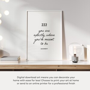 May include: A black and white printable art print with the text "222 you are exactly where you're meant to be ALIGNMENT". The print is framed and sitting on a wooden surface with a lamp.