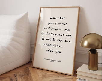 May include: A framed print with the lyrics "now that you're mine, we'll find a way of chasing the sun, let me be the one that shines with you." The print is in a wooden frame and is leaning against a wall. A gold lamp and books are next to the frame.