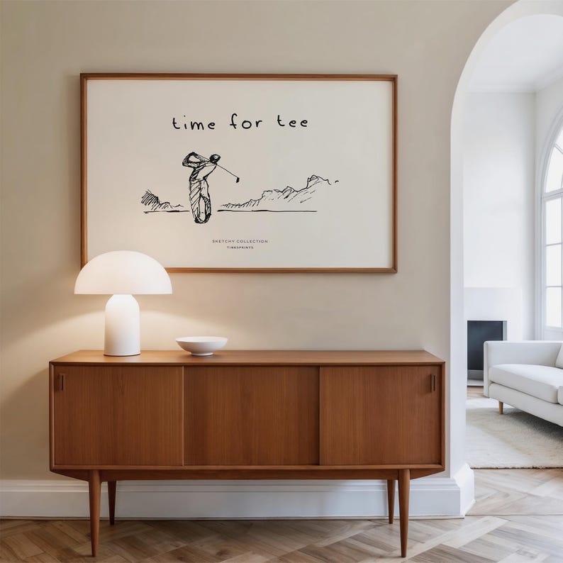 May include: A framed print with the text "time for tee" and a sketch of a golfer swinging. The print hangs above a mid-century modern wooden cabinet with a white lamp and bowl. The room has a neutral color palette.