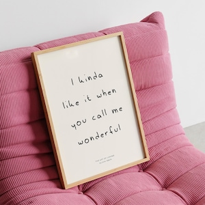 May include: A framed print with the text "I kinda like it when you call me wonderful" in black script. The print has a light wood frame and is set against a pink corduroy upholstered chair. The bottom of the print reads "THE ART OF LOVING OLIVIA DEAN".
