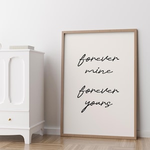 May include: A framed print with the words "forever mine forever yours" in black script on a white background. The frame is a light brown colour. The print is leaning against a white wall.