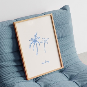 May include: A framed art print featuring two blue palm trees on a white background. The print has the words "easy breezy" in a handwritten font. The frame is made of light-colored wood. The artwork is displayed on a blue corduroy chair.