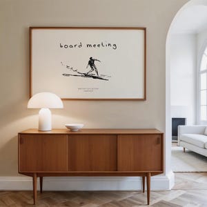 May include: A mid-century modern style wooden cabinet with sliding doors and tapered legs. A white table lamp with a rounded shade and a small white bowl sit on top. A framed print with the words "board meeting" and a surfer illustration hangs above.