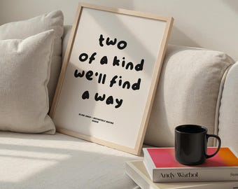 May include: A framed print with the text "two of a kind we'll find a way" in black on an off-white background. The print is in a light wood frame. A black mug sits on a stack of books, including one by Andy Warhol.