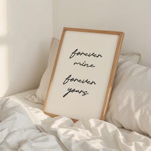 May include: A rectangular framed print with the words "forever mine forever yours" in black script. The print is in a light wood frame, set against white bedding. The image has a minimalist and romantic feel.