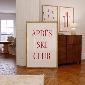 May include: A framed print with the words "APRÈS SKI CLUB" in a bold, red font. Two additional framed prints feature skiers and red skis. The prints are displayed in a room with a wooden floor and a wooden cabinet.