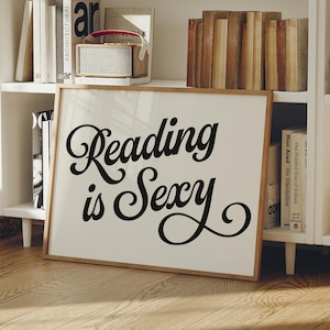 May include: A framed print with the words "Reading is Sexy" in elegant black script. The artwork has a light background and a natural wood frame. Bookshelves with books and a small radio are visible in the background.