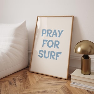 May include: A framed print with the text "PRAY FOR SURF" in blue lettering on a white background. The frame is light wood.