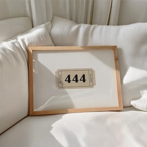 May include: A framed print featuring a beige ticket with the number "444" in bold black font. The ticket is centered on a white background, within a light brown wooden frame. The words "ANGEL NO." are printed vertically on both sides of the ticket.