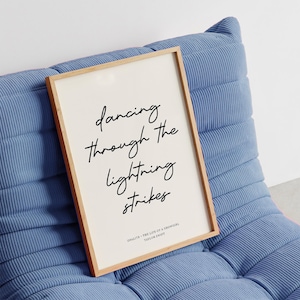 May include: A framed print with the words "dancing through the lightning strikes" in black script. The print has a light wood frame and a cream-coloured background. The print is resting on a blue corduroy chair.