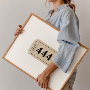 Könnte beinhalten: Gerahmtes Kunstwerk mit einem beigen Ticket-Design, das die Zahl "444" in Schwarz zeigt. Das Ticket ist vor einem weißen Hintergrund platziert und von einem hellbraunen Holzrahmen umgeben. Die Worte "ANGEL NO." sind auf dem Ticket aufgedruckt.