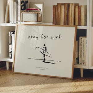 May include: A framed print with the words "pray for surf" above a black and white illustration of a person holding a surfboard. The print has a light wood frame and is displayed on a white shelf with books.