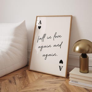 Op de afbeelding: Een ingelijste print met de tekst "fall in love again and again" in zwart schrift. De print toont het harten aas kaart symbool aan de boven- en onderkant. Een goudkleurige lamp en boeken zijn zichtbaar op de achtergrond.