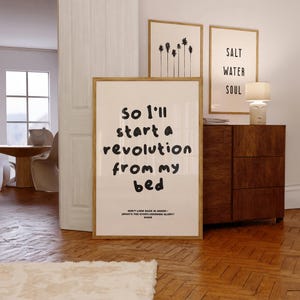 May include: A framed art print with the text "So I'll start a revolution from my bed" in a modern font. The print is in a room with other framed art and a wooden cabinet. The text is a quote from the Oasis song "Don't Look Back in Anger."