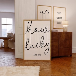 Op de afbeelding: Een ingelijst print met de tekst "how lucky are we" in zwarte cursieve letters op een witte achtergrond. De print hangt aan een witte muur naast twee andere ingelijste prints.