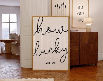 May include: A framed print with the text "how lucky are we" in black cursive script on a white background. The print is hanging on a white wall next to two other framed prints.