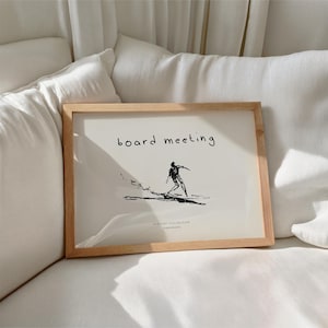 May include: A framed print with a light wood frame. The print features a black and white illustration of a person surfing, with the words "board meeting" above the illustration. The text "SKETCHY COLLECTION" and "TIMESPRINTS" are below the illustration.