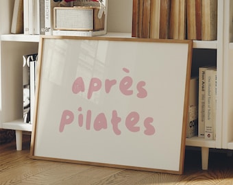 Puede incluir: Una impresión enmarcada con las palabras "après pilates" en letras rosas de burbujas. La impresión tiene un fondo beige claro y un marco de madera natural. La impresión se muestra en una estantería blanca con libros y una pequeña radio.