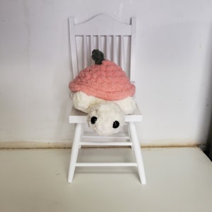 Puede incluir: Una tortuga de peluche hecha a mano con caparazón rosa y cuerpo blanco, sentada en una pequeña silla blanca. La tortuga tiene ojos negros grandes y un pequeño tallo verde en su caparazón. El peluche está hecho de hilo suave y texturizado.