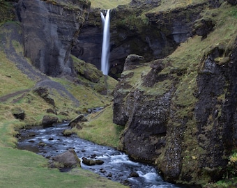 Könnte beinhalten: Ein Wasserfall stürzt von einer felsigen Klippe herab und bildet einen kleinen Bach, der sich durch ein grünes Tal schlängelt.