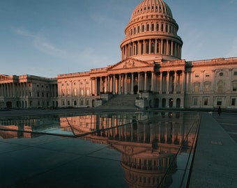 May include: A view of the United States Capitol Building in Washington, D.C. The building is made of white stone and has a large dome. The building is reflected in a large, rectangular pool of water in front of it.