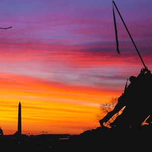May include: Silhouette of the Iwo Jima Memorial against a vibrant sunset sky. The sky is a mix of orange, pink, and purple hues. The Washington Monument is visible in the distance.