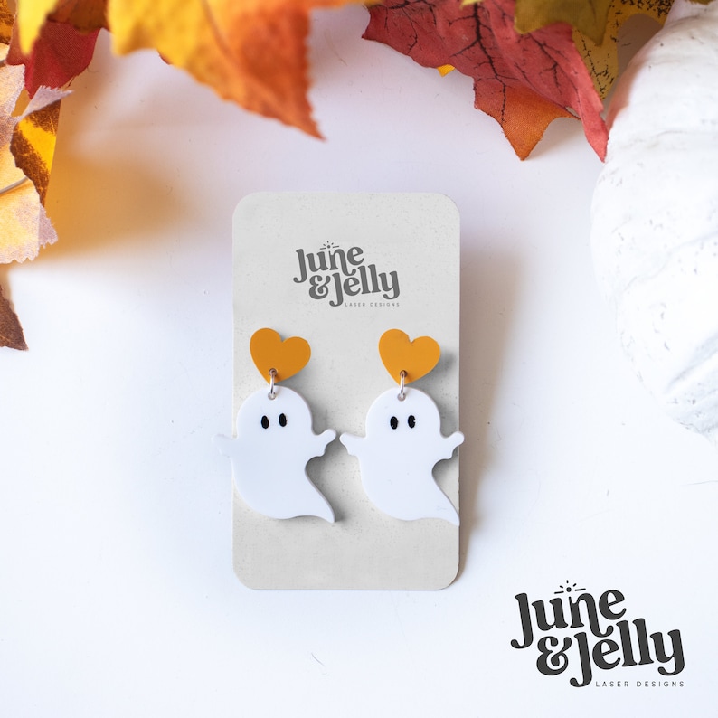 May include: White ghost shaped earrings with a small yellow heart charm. The earrings are on a white card with the text "June & Jelly Laser Designs".