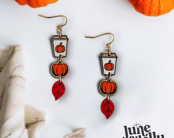 May include: A pair of dangle earrings featuring a fall-themed design. Each earring has a small coffee cup with a pumpkin inside, a pumpkin charm, and a red leaf charm. The earrings are made of wood and have gold hooks.