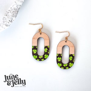 May include: A pair of wooden earrings with a green shamrock pattern on a black background. The earrings are oval shaped with a gold hook.