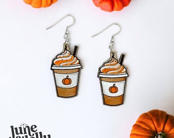 May include: A pair of dangle earrings featuring a pumpkin spice latte design. The earrings are made of wood and painted with a realistic depiction of a coffee cup with whipped cream and a sprinkle of cinnamon. The earrings are a fun and festive way to show your love for fall.