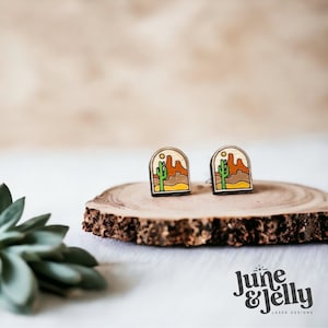 Op de afbeelding: Een paar houten oorbellen met een woestijnlandschap ontwerp. De oorbellen zijn geschilderd in tinten bruin, oranje, geel en groen. Het ontwerp bevat een cactus, bergen en een zon.