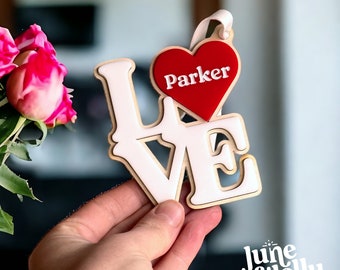 May include: White wooden "LOVE" sign with a red heart that says "Parker" on it. The sign is being held by a hand. The sign is a laser cut design from June & Jelly Laser Designs.