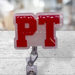 May include: A badge reel with a rectangular, glittery white base. The letters "PT" are in bold, red font. The badge reel has a silver-colored clip and a retractable cord.