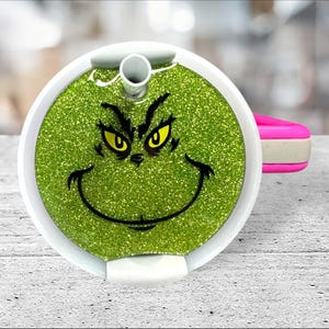 May include: A white and green glitter tumbler lid with a Grinch face design. The lid has a straw hole and a pink handle.