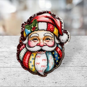 Puede incluir: Adorno de cara de Papá Noel colorido con un gorro rojo y blanco, barba blanca y expresión sonriente. La barba y el gorro tienen un diseño de patchwork con varios patrones y colores. Una ramita de acebo adorna el gorro.