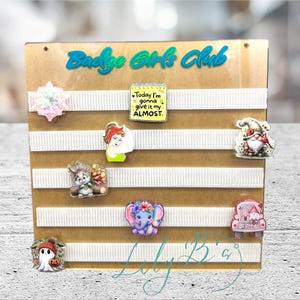 Op de afbeelding: Een displaybord met de tekst "Badge Girls Club" in blauw en diverse decoratieve badge-spelden. De spelden bevatten een sneeuwvlok, een kabouter, een cartoon-olifant, een spook, een auto, een vrouw en een speld met de tekst "Today I'm gonna give it my ALMOST."