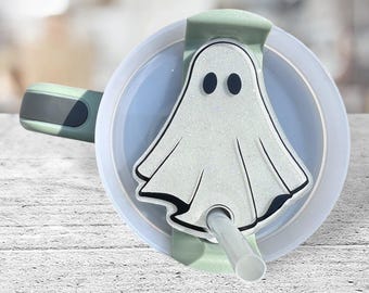 May include: A white ghost-shaped lid with glitter accents and black eyes, attached to a green lid for a tumbler. The lid has a hole for a straw.