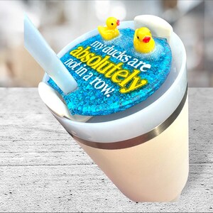 May include: A white and beige tumbler with a blue glitter lid featuring yellow text that reads "my ducks are absolutely not in a row." Two yellow rubber duckies float on the blue glitter. A white straw is inserted into the tumbler.