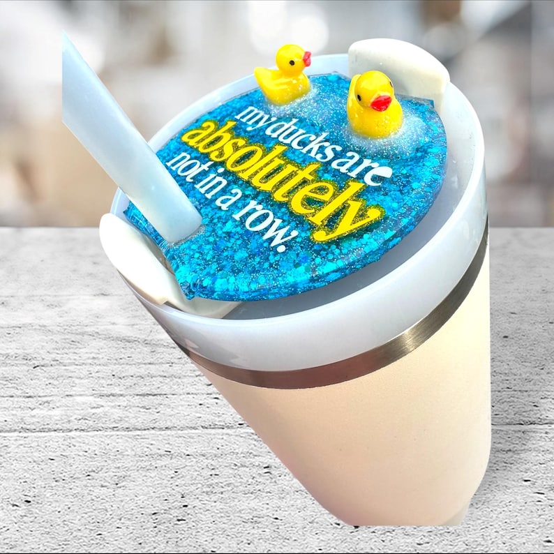 May include: A white and beige tumbler with a blue glitter lid featuring yellow text that reads "my ducks are absolutely not in a row." Two yellow rubber duckies float on the blue glitter. A white straw is inserted into the tumbler.