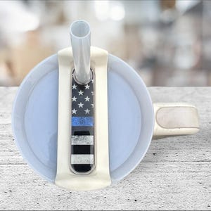 May include: A white travel mug with a blue and white American flag with a thin blue line design on the lid. The mug has a clear plastic straw.