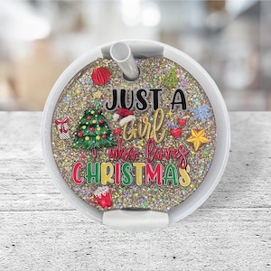 May include: A round, white glitter tumbler with a straw. The tumbler features the text "JUST A Girl who loves CHRISTMAS" with Christmas-themed graphics, including a Christmas tree, ornaments, and a Santa hat.