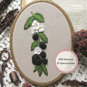 Peut inclure: Un tambour à broder ovale en bois contenant un panneau en tissu avec un motif de ronces de mûres cousu à la main. La broderie présente des fleurs blanches à cœur jaune, des feuilles vertes et des mûres sombres texturées. Une étiquette rose circulaire indique PDF Pattern & Instructions.