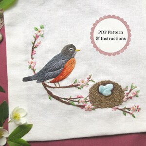 May include: Embroidered design of a robin on a branch with a nest and pink blossoms. The bird has grey and orange feathers. The text "PDF Pattern & Instructions" is in a pink circle.