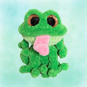 May include: A plush green frog with large, glittery gold-rimmed eyes and a pink tongue. The frog is made of a soft, textured material. The frog's body is a vibrant green, with a lighter green on its belly and a pink tongue.