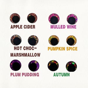 May include: A collection of six pairs of decorative doll eyes with glitter accents. Each pair has a black pupil and a colored iris, with text labels: Apple Cider, Mulled Wine, Hot Choc-Marshmallow, Pumpkin Spice, Plum Pudding, and Autumn.